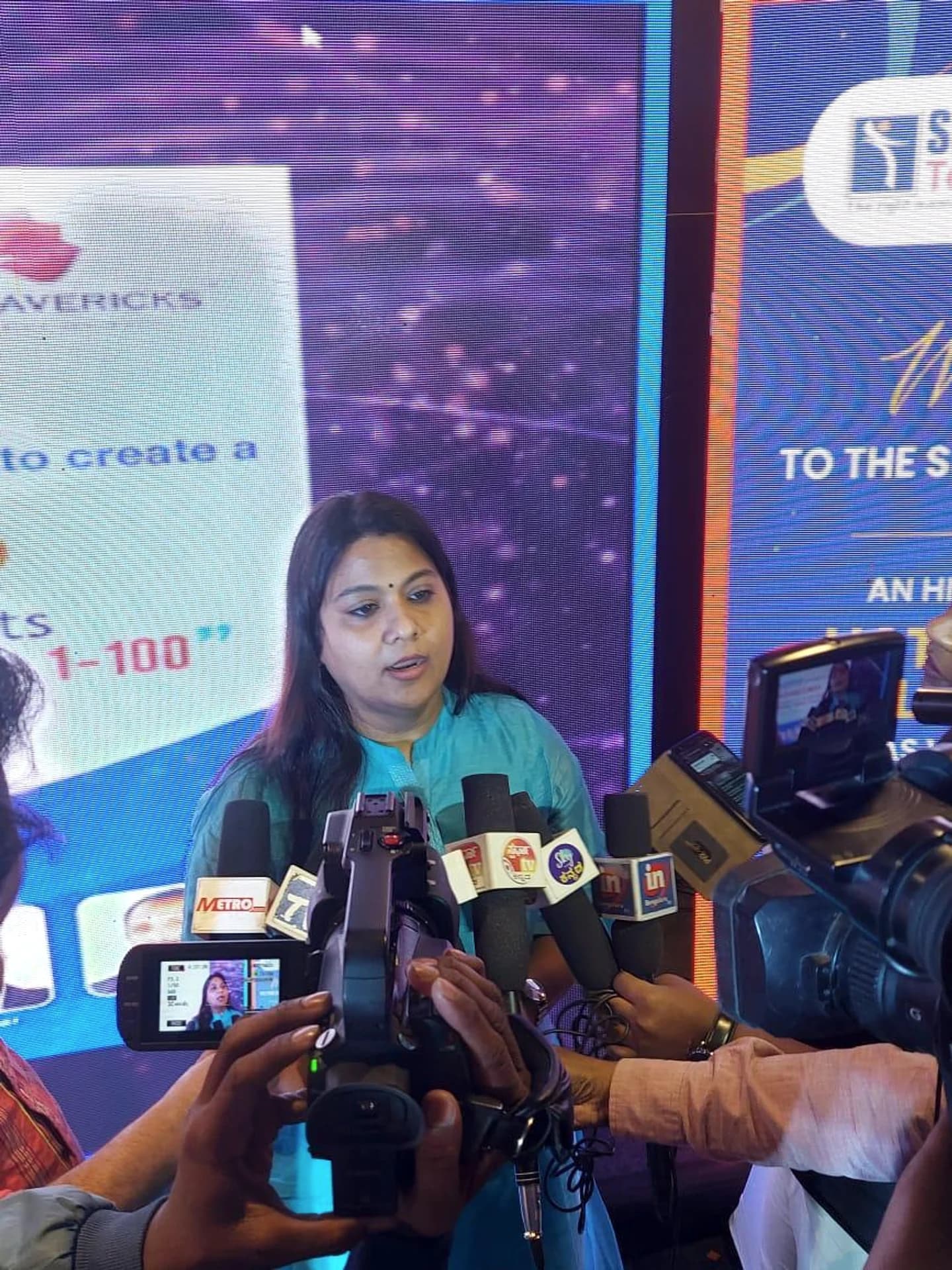 Pushpavalli speaking with journalists, surrounded by microphones and cameras at a Mavericks event.