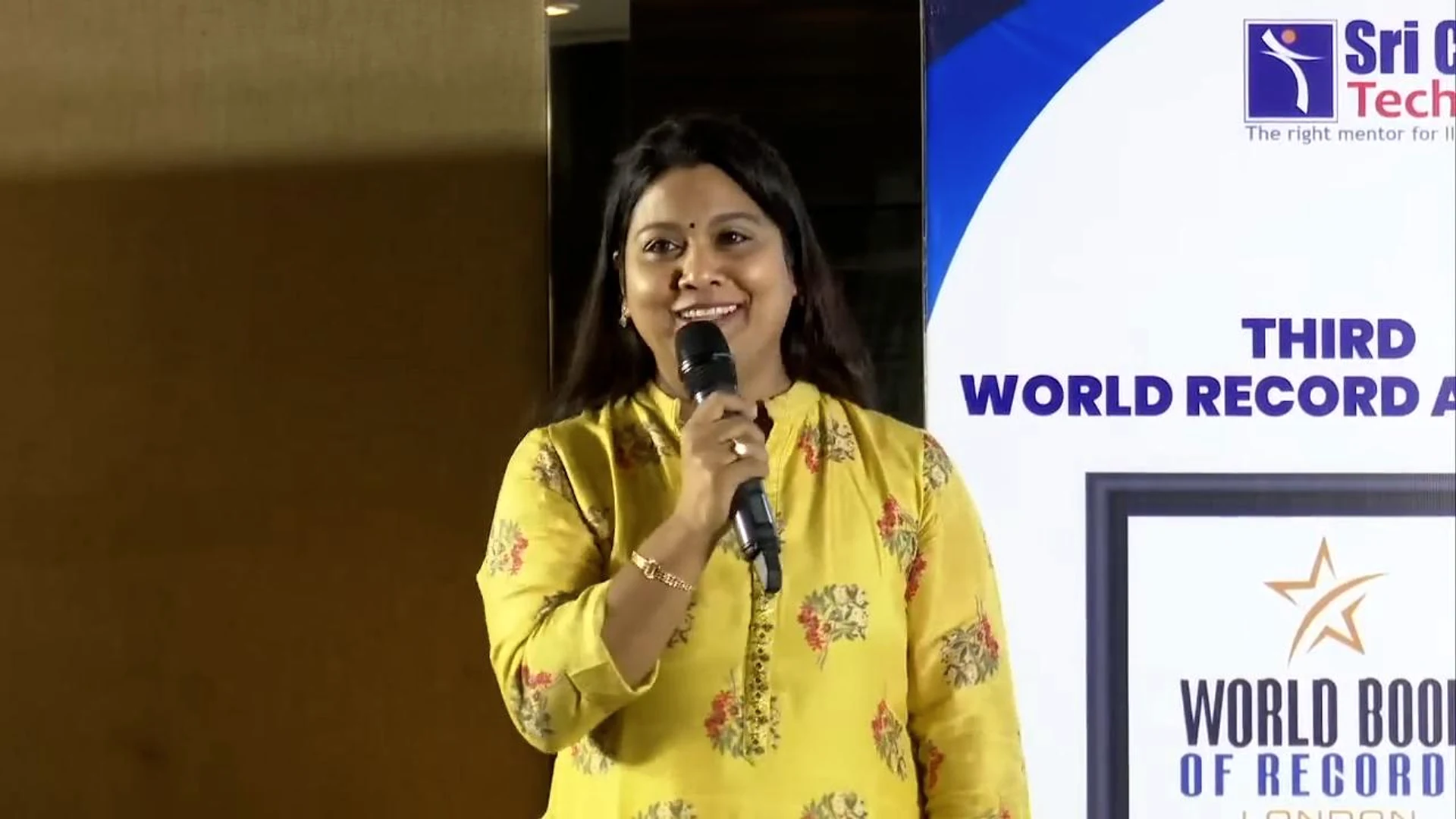 Pushpavalli speaking beside a world record event banner.
