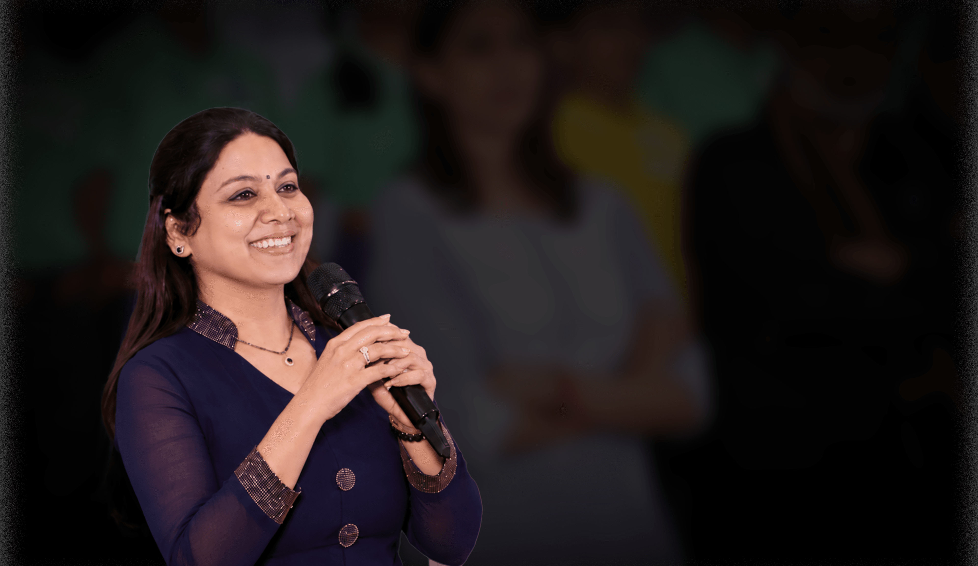 Pushpavalli smiling and speaking into a microphone against a softly blurred event backdrop.
